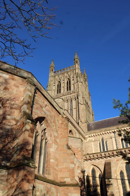 The view is looking up at the tower in pale brown stone. In the foreground is the edge of the chapter house wall and in the middle distance is the exterior of the quire. The sky s a cloudless blue colour.