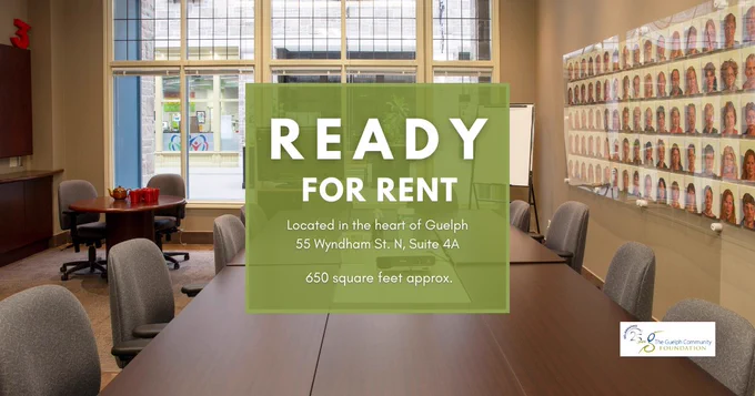 Boardroom with a long meeting table, chairs, tv, projector, small table with two chairs, and big windows at The Guelph Community Foundation office. And a big sign in the middle that says "Ready for rent, located in the heart of Guelph, 55 Wyndham St, N, Suite 4A, 650 square feet approx."