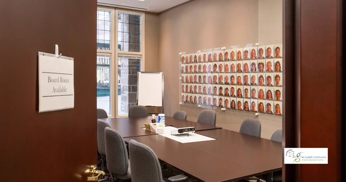 Boardroom with a long table and chairs, open door with a “Board Room Available” sign, wall of portraits, and Guelph Community Foundation logo visible