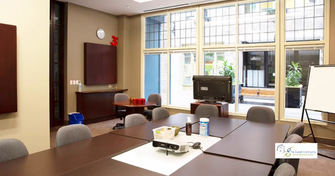 Bright boardroom with large windows, long meeting table, chairs, TV screen, recycle blue bin, clock in the wall, whiteboard, and Guelph Community Foundation logo visible