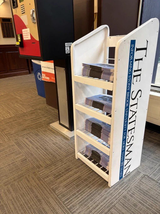 White rack filled with stacks of The Statesman newspapers inside a building lobby.