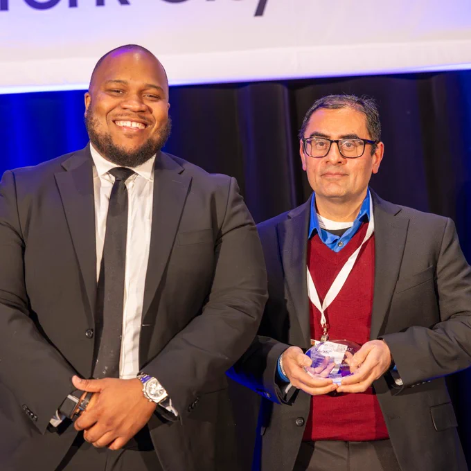 Two people in suits stand smiling on stage, one holding an award during a recognition ceremony.