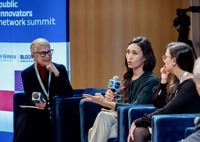 Panelists speak at a public innovators network summit with a Johns Hopkins University banner in the background. One participant holds a microphone.