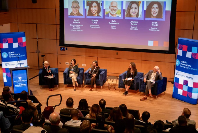 Panel discussion at the Public Innovators Network Summit with five speakers seated on stage, each with name and affiliation displayed above them. Audience in foreground, looking towards the stage.