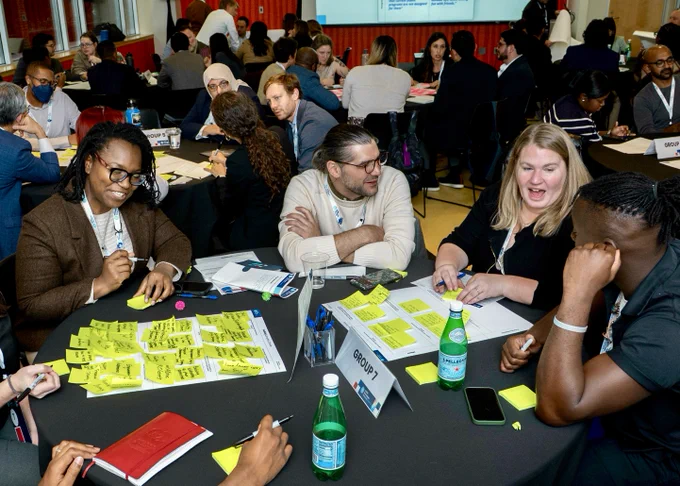 Group of people engaged in a workshop, sitting at tables with papers and post-it notes, discussing ideas in a lively manner.