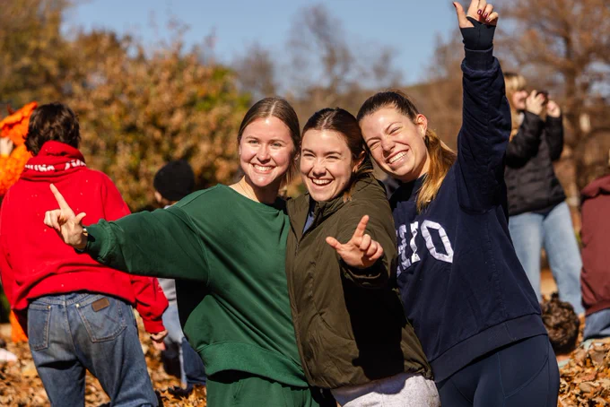 Three smiling individuals, dressed in casual attire, enjoying an outdoor autumn setting.