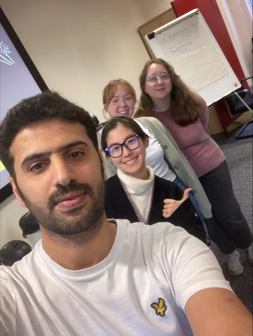 Four people posing for a group selfie in a casual office setting with a flip chart and projector screen in the background.
