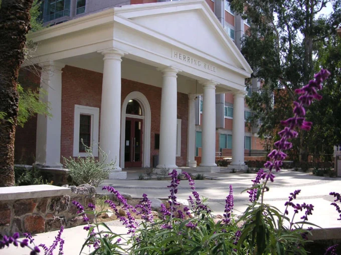 Herring Hall. A small Roman Revival building with a prostyle portico and arched entrance with purple flowers in the foreground.