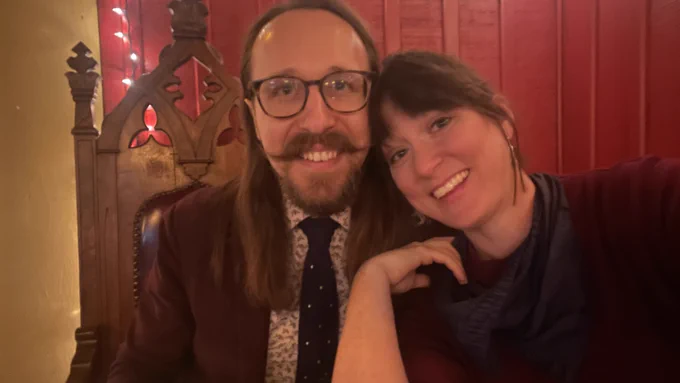 Me and my partner Sarah. I'm in the throne-like chair at The Publick House in a burgundy suit, brown tortoiseshell glasses, brown hair past shoulder length, and a beard with a long moustache. Sarah leans in from the viewer's right in a burgundy sweater with a gray(?) scarf. She has brown hair with bangs. We're smiling wide.