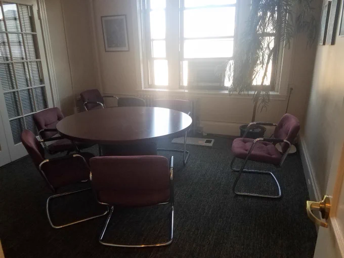 A round table with five upholstered metal chairs. One chair sits separate, facing the rest. Light pours in from a window opposite the camera. This is the room in which my defense took place, photographed two hours before the defense began.