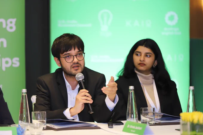 A young man in a black suit and glasses speaks into a microphone at a roundtable discussion, gesturing with one hand, while a woman beside him listens attentively; green branding for The Quantum Hub, Duolingo English Test and Kaio Communications forms the backdrop, highlighting a conversation on digital, AI-enabled assessment in India.