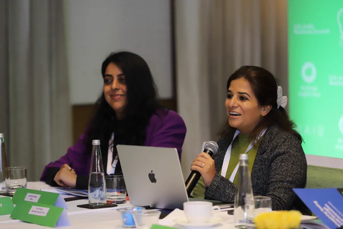 Two women sit at a conference table with laptops, microphones, name placards and water glasses; one in a purple blazer and one in a grey jacket and green top, smiling as she speaks, against a green backdrop referencing Duolingo English Test and partners at a roundtable on reimagining testing in India.