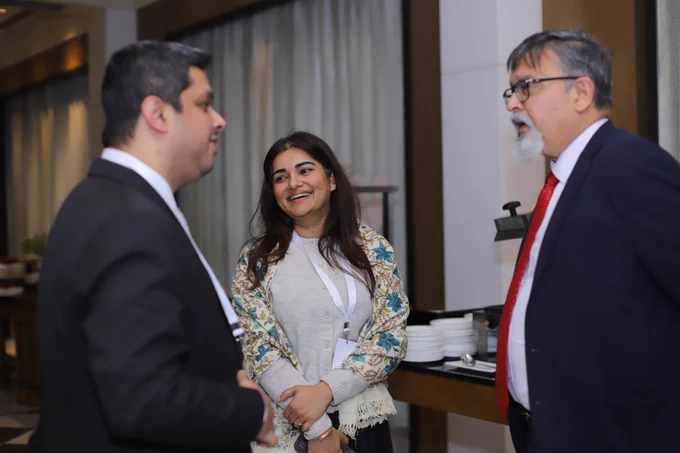 Three professionals network informally during the event coffee break; a woman in a light cardigan and floral shawl smiles while conversing with two men in suits and lanyards, standing near a buffet setup, capturing the collaborative spirit behind modernising India’s testing ecosystem.