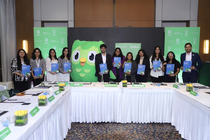 A group photo of the roundtable participants shows ten professionals standing in front of event branding and a large Duolingo owl mascot, each holding copies of the “Reimagining Testing in India through Digital Innovation” white paper, symbolising a shared commitment to expanding access to higher education through secure, AI-enabled digital testing.
