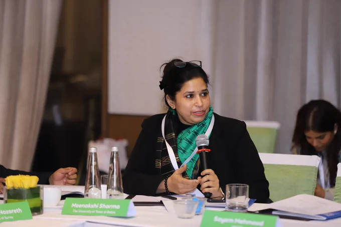 A woman in a black blazer and patterned green scarf speaks into a microphone during the roundtable, seated at a conference table with name placards, notebooks, and water glasses, reflecting an engaged discussion on reimagining India’s testing and higher education ecosystem.