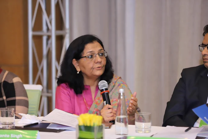 A woman in a bright pink kurta and embroidered shawl speaks into a microphone at the roundtable, gesturing with one hand while seated at a conference table scattered with papers, notepads, and water glasses, highlighting active dialogue on reimagining testing and access to higher education in India.