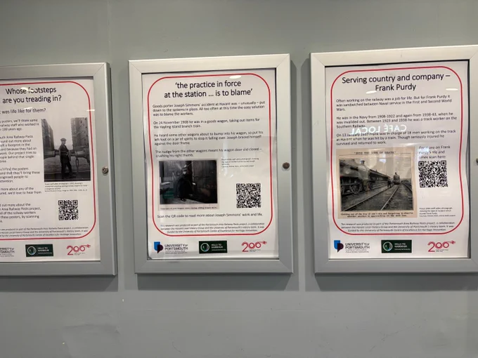 Three posters in frames on a station ticket office wall. One is a general introduction; the others detail the life stories of Frank Purdy and Joseph Simmons, including posed staff safety photographs, text and QR codes in a red border, with logos of the University of Portsmouth Centre of Excellence for Heritage Innovation, Hills to Harbour CRP and Railway 200.