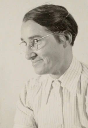 Black and white portrait of a woman smiling, wearing round glasses and a striped shirt.