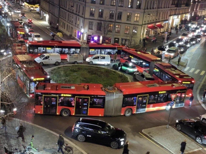 Multiple articulated red buses jammed in a circular roundabout with cars, vans and pedestrians navigating the crowded downtown intersection at night