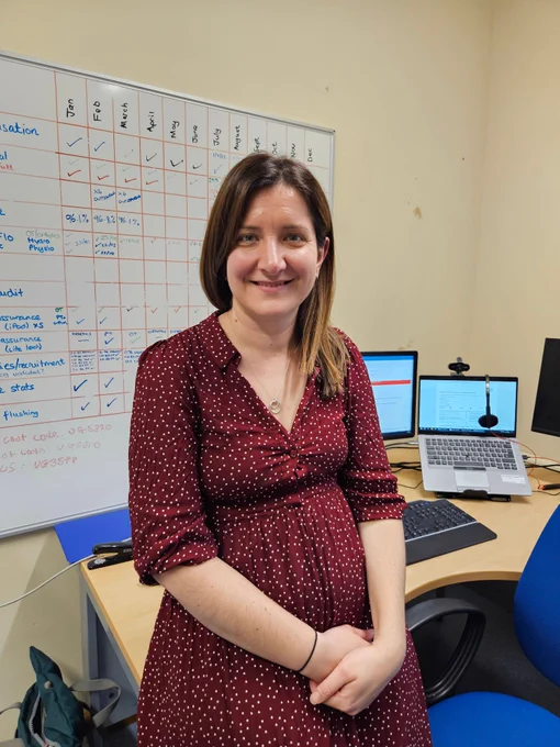 Woman wearing a maroon dress with white dots posing in a workspace featuring a whiteboard filled with notes and a laptop on the desk.