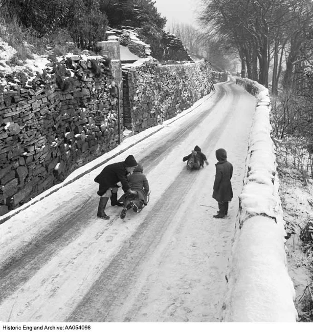 Black and white photograph of a group of four children playing with sledges along a narrow, snowy road bounded by stone walls. One child watches as another slides down the road. Next to them, a child pushes a sledge with another child lying on it.