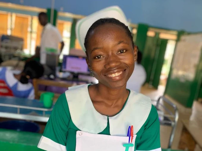 A healthcare worker in green scrubs and a badge smiles warmly in a clinic setting, with medical equipment and another worker in the background.