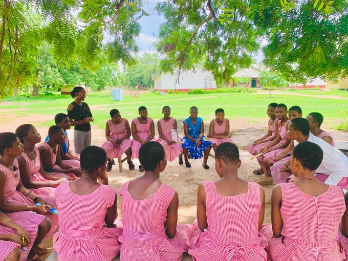 A group of students in matching pink uniforms seated in a circle on the ground outdoors, engaging in a discussion led by an instructor standing to the side, surrounded by lush greenery.