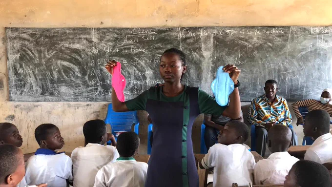 A teacher demonstrating the use of sanitary pads to students in a classroom, with a chalkboard in the background filled with writing.