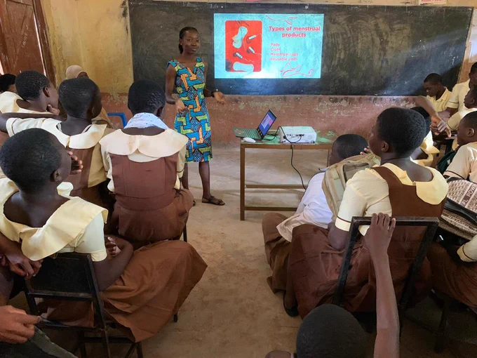 Josephine presents a slide about types of menstrual products to a classroom of students in uniforms, with a laptop visible on a desk and a blackboard in the background.