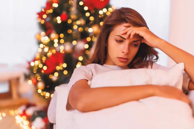 Person looking thoughtful while sitting on a sofa with a Christmas tree lit up in the background.