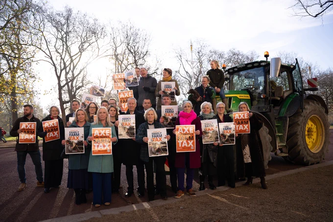 A group of Lib Dem MPs standing together on the Mall, holding orange and white protest signs that read “AXE THE FAMILY FARM TAX.” They pose in front of leafless trees on a road, with a green tractor parked behind them. Some people wear coats and scarves.