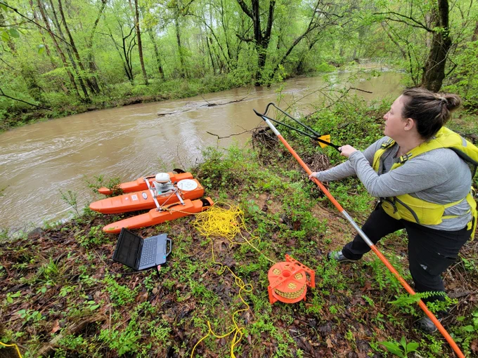 A Woman on the bank of a stream using a slingshot to get equipment to the other side of the stream.