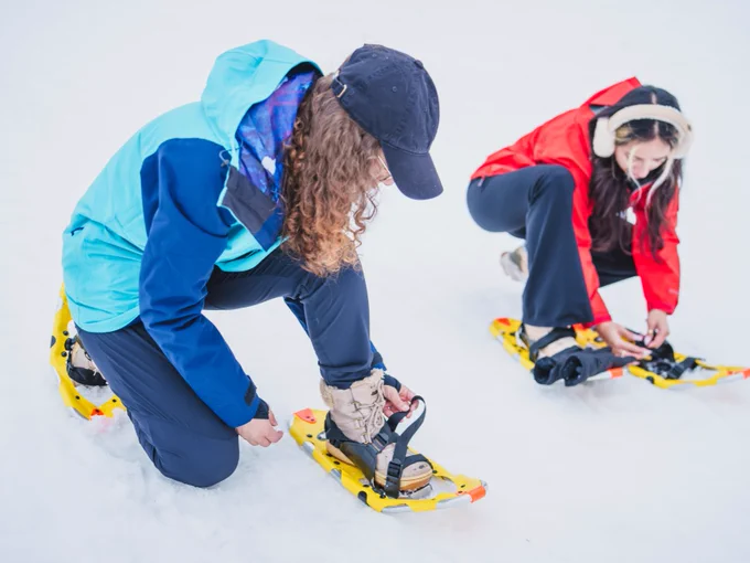 Two individuals wearing colorful winter gear are adjusting their snowshoes on a snowy ground.