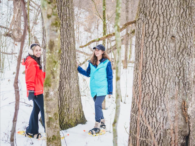Two people wearing snowshoes are smiling and chatting in a snowy forest. One individual is dressed in a red jacket and the other in a blue jacket. Both are standing near trees.