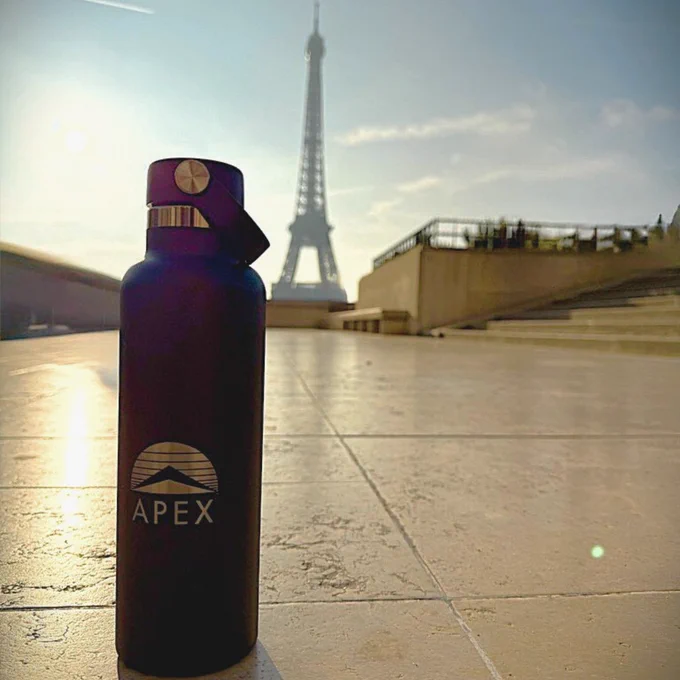 A navy blue Apex Systems water bottle in focus on a paved surface, with the Eiffel Tower in the background under a clear sky.