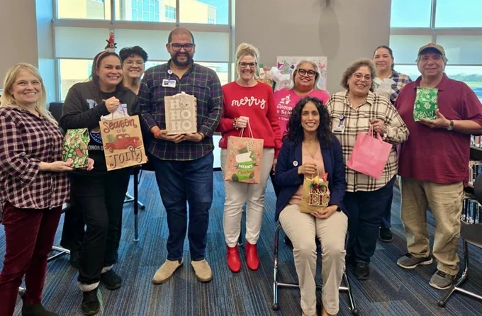 ⸻  Alt Text – Image 1: Large group of smiling staff members in a school library holding books and holiday gift items during a festive Hug in a Mug and book exchange event.