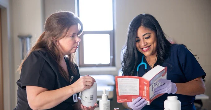 Two healthcare professionals are engaged in a discussion over a book titled "Merck Manual" in a clinical setting, surrounded by medical supplies.