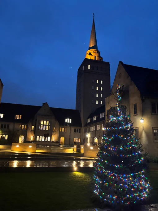 Nuffield College at twilight with a Christmas tree in the foreground. The Library tower and other college buildings are lit with warm yellow lights against a rich blue twilight sky. The ground glitters with rain, and in the foreground is a Christmas tree with multicoloured lights.