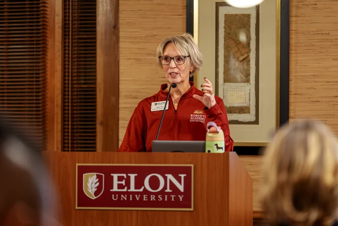 A person is giving a speech at a podium with the Elon University logo and microphone. They are gesturing with one hand.