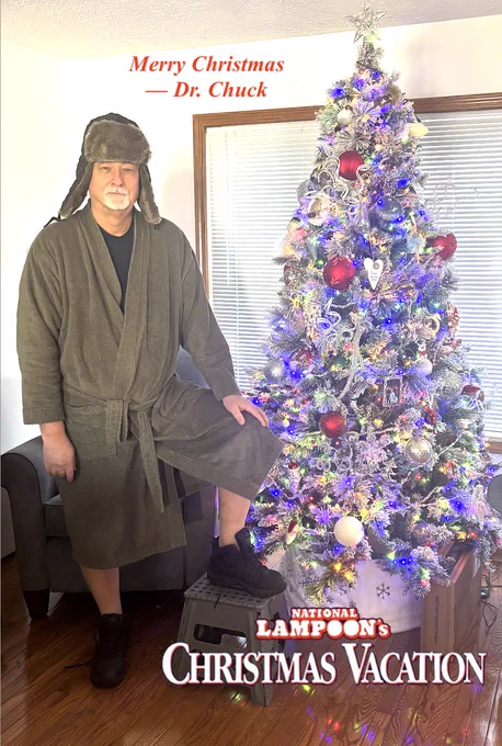 Dr Chuck reenacting a costume from Christmas Vacation with a ratty old olive drab robe and untied shoes and an Alaskan fur hat standing next to a Christmas Tree.