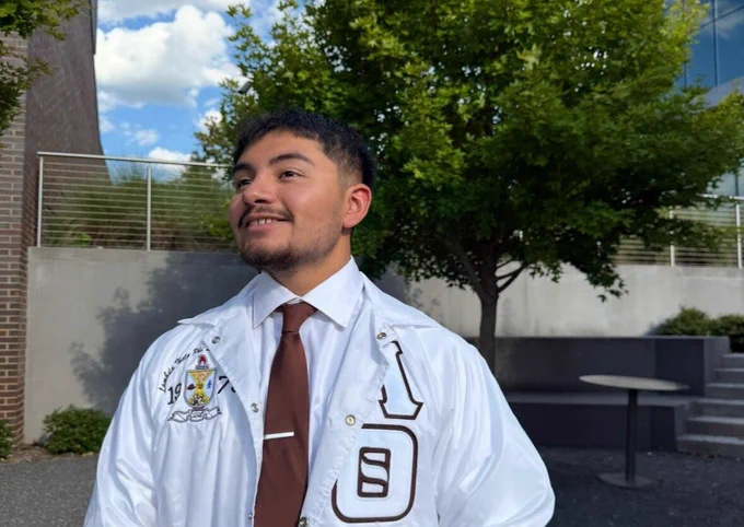 Person wearing a white jacket with embroidered patches and a brown tie standing outdoors near a tree and modern building.