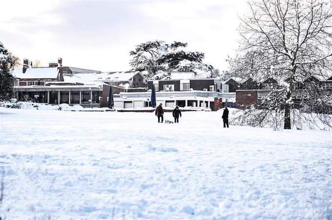 Three people walk across a snow-covered field toward a University of Reading building with snow on its roof and surrounding trees.