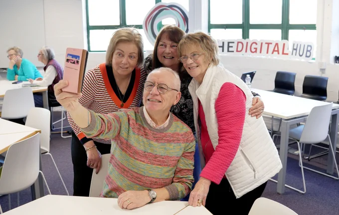 A person using a smartphone to take a selfie with three other people. The person holding the phone is sitting down at a table while the other three people are standing. They are all smiling at the camera while posing for the photo.