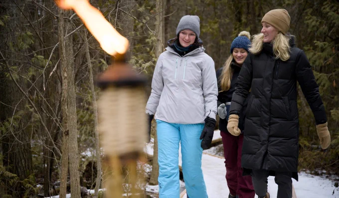 Three individuals walking along a snowy forest path, smiling and dressed in winter clothing, with a lit torch in the foreground.