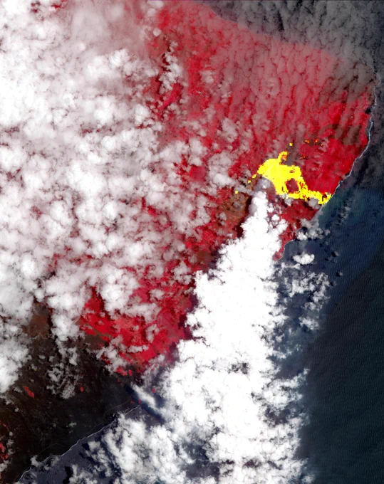 Satellite view showing a volcanic eruption with bright yellow lava flow and surrounding areas of vegetation depicted in red under scattered clouds by the coastline.