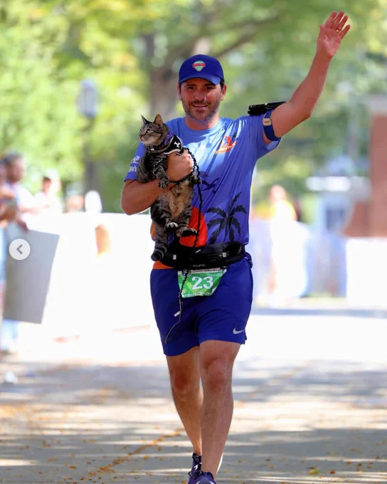 A man wearing blue athletic gear and wearing the number 23 near the finish line of a race. He's holding a gray tabby one arm and waving with the other.