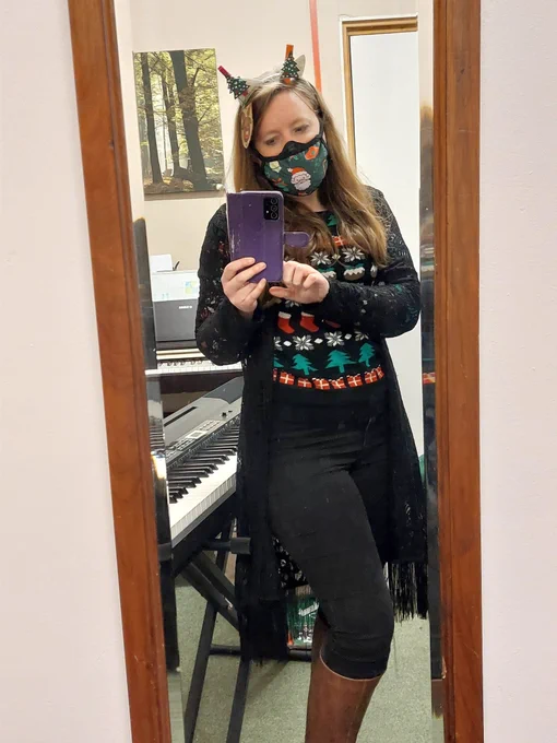 A mirror selfie I'm wearing a black Christmas jumper with red stockings, white snowflakes, Christmas puddings, christmas trees and red presents. I'm wearing a black ffp3 mask with a Christmas mask with presents and father Christmas. I'm wearing silver ears with Christmas trees on top, and a rudolf decoration made by one of my students. 2 keyboards are visible behind me in my teaching room. 