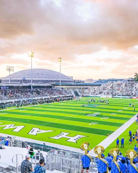 View of a football game at University of Hawaii Stadium, with a marching band performing in the foreground and fans in the stands under a cloudy sky.