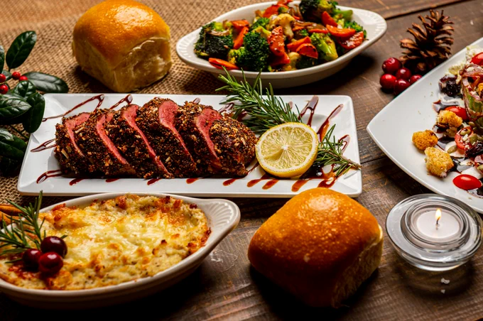 Holiday feast on a wooden table featuring sliced meat with lemon and rosemary, cheesy potatoes, roasted vegetables, salad, rolls, a candle, and festive decorations, creating a warm and inviting ambiance.