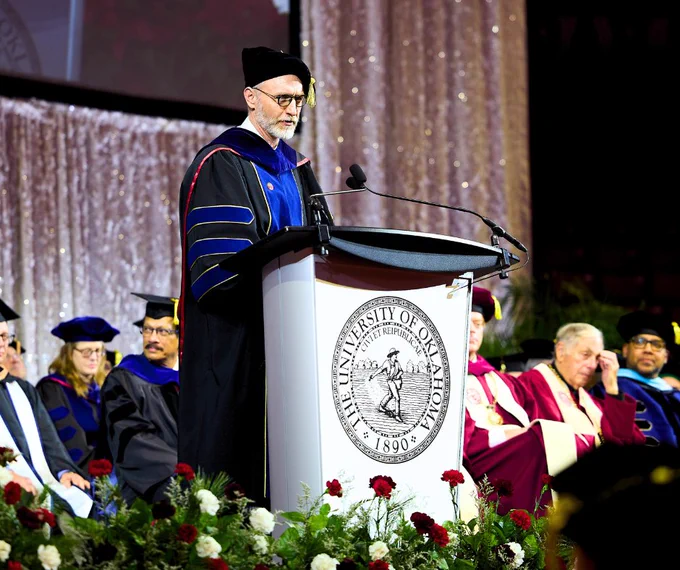 Person in academic regalia delivering a speech at a University of Oklahoma podium during a graduation ceremony, with faculty members seated in the background.